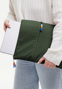 Sacca per laptop verde imbottita con doppia zip e linguette colorate. Texture liscia, progettata per adattarsi a un laptop, tenuta da una persona.