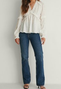 Blusa bianca con texture e dettagli arricciati, scollatura a V e maniche lunghe abbinata a jeans blu a zampa d'elefante. Sandali neri con cinturini completano il look.