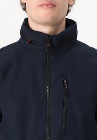Giacca blu navy con colletto alto, zip frontale e due tasche con zip. Tessuto liscio con un aspetto testurizzato e accenti in metallo a contrasto.