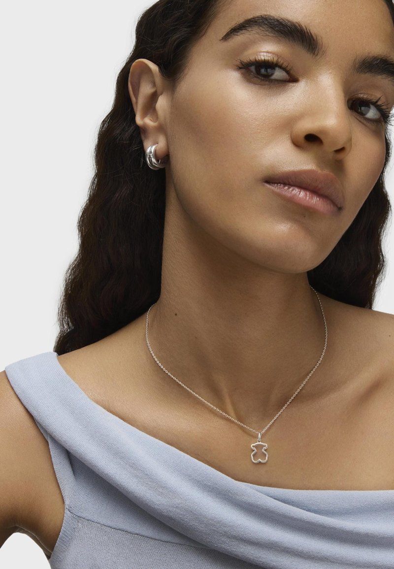 Srebrny naszyjnik w kształcie misia na cienkim łańcuszku, w połączeniu z dużymi srebrnymi kolczykami w kształcie kół, noszonym z miękką niebieską bluzką bez ramiączek.