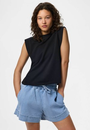 Frau mit lockigem Haar, die ein ärmelloses schwarzes Oberteil und hellblaue Denim-Shorts mit elastischem Bund trägt, Hände in den Taschen.