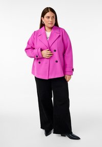 Dubbelknäppt rosa kappa med breda slag och knappdetaljer, i kombination med svarta vida byxor och svarta spetsiga skor.