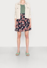 Nainen, jolla on päällä vihreä neulepaita, kerma valkoinen takki, tummansininen kukallinen volangihame ja vihreät punotut korkokengät, seisoo yksinkertaisella harmaalla lattiapinnalla.