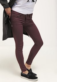 Jeans slim fit burdeos con una textura suave, que cuentan con bolsillos frontales, cremallera y un diseño que se estrecha hacia el tobillo. Combinados con zapatos negros tipo slip-on.