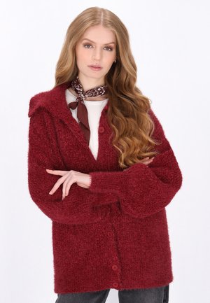 Bordeaux kleurige chunky gebreide cardigan met een kraag, grote knopen en wijde mouwen. Draagt over een wit topje, aangevuld met een bruine patroon sjaal.