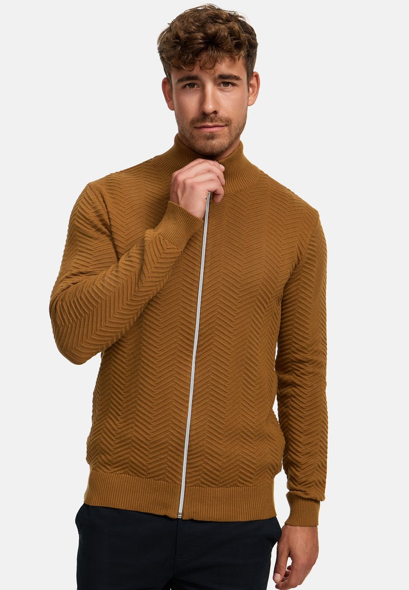 Brun zip-up tröja med ett strukturerat chevronmönster, hög krage, ribbade ärmslut och en kontrasterande dragkedja. Bärs över mörka byxor.