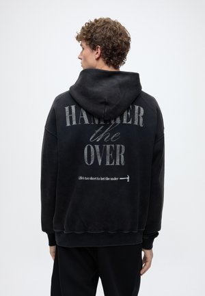 Млад мъж с къдрави коси, облечен в черно суичър с качулка с текст "HAMMER the OVER" и малка графика на чук на гърба, на едноцветен фон.