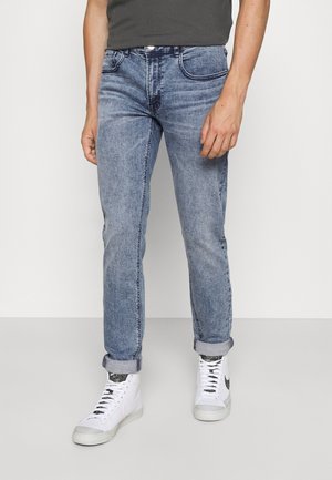 Vaqueros de mezclilla azul claro con corte recto, bolsillos frontales y puños doblados. Llevados con zapatillas blancas con detalles en gris.