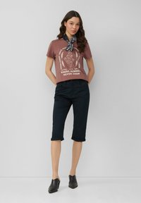 Kurzärmliges burgunderrotes T-Shirt mit weißem grafikdesign; schwarze, cropped Hosen; schwarze Slipper; geknoteter schwarz-weißer Schal um den Hals.