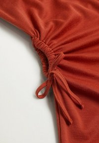 Tissu couleur rouille avec des détails froncés rassemblés par un cordon noué sur un fond blanc lisse.