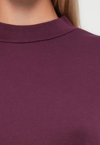 Gros plan sur une personne portant un pull col roulé violet, avec de longs cheveux blonds visibles sur le côté.