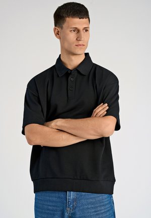 Schwarzes Polohemd mit drei Knöpfen und Kragen, mit kurzen Ärmeln und lockerer Passform, getragen mit blauen Jeans.