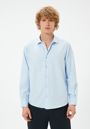 Camicia a maniche lunghe azzurro chiaro con colletto a punta, chiusura con bottoni e orlo curvo. Il tessuto è liscio con una leggera lucentezza. Senza fantasie.