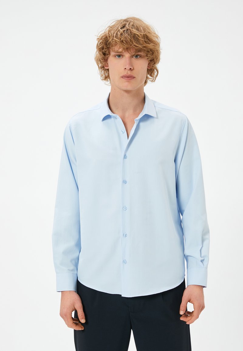 Camicia a maniche lunghe azzurro chiaro con colletto a punta, chiusura con bottoni e orlo curvo. Il tessuto è liscio con una leggera lucentezza. Senza fantasie.