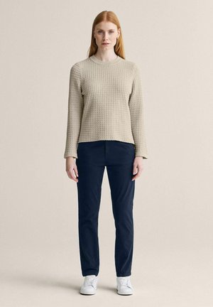 Kvinde med glat rødt hår står vendt fremad iført en beige tekstureret langærmet sweater, mørkeblå bukser og hvide sneakers.