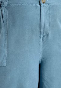 Light blue pants made of soft fabric, featuring a button closure, visible stitching, and a side pocket. Smooth texture, casual design.