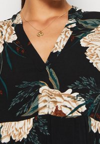 Mujer con un vestido negro de flores y escote en V, y un collar de oro con un colgante circular.