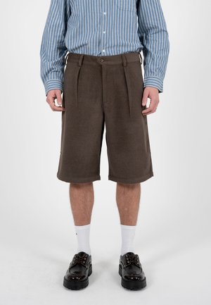 Mężczyzna ubrany w brązowe krótkie spodenki do kolan, niebiesko-białą pasiastą koszulę z długim rękawem, białe skarpetki i czarne masywne buty stoi na białej podłodze.