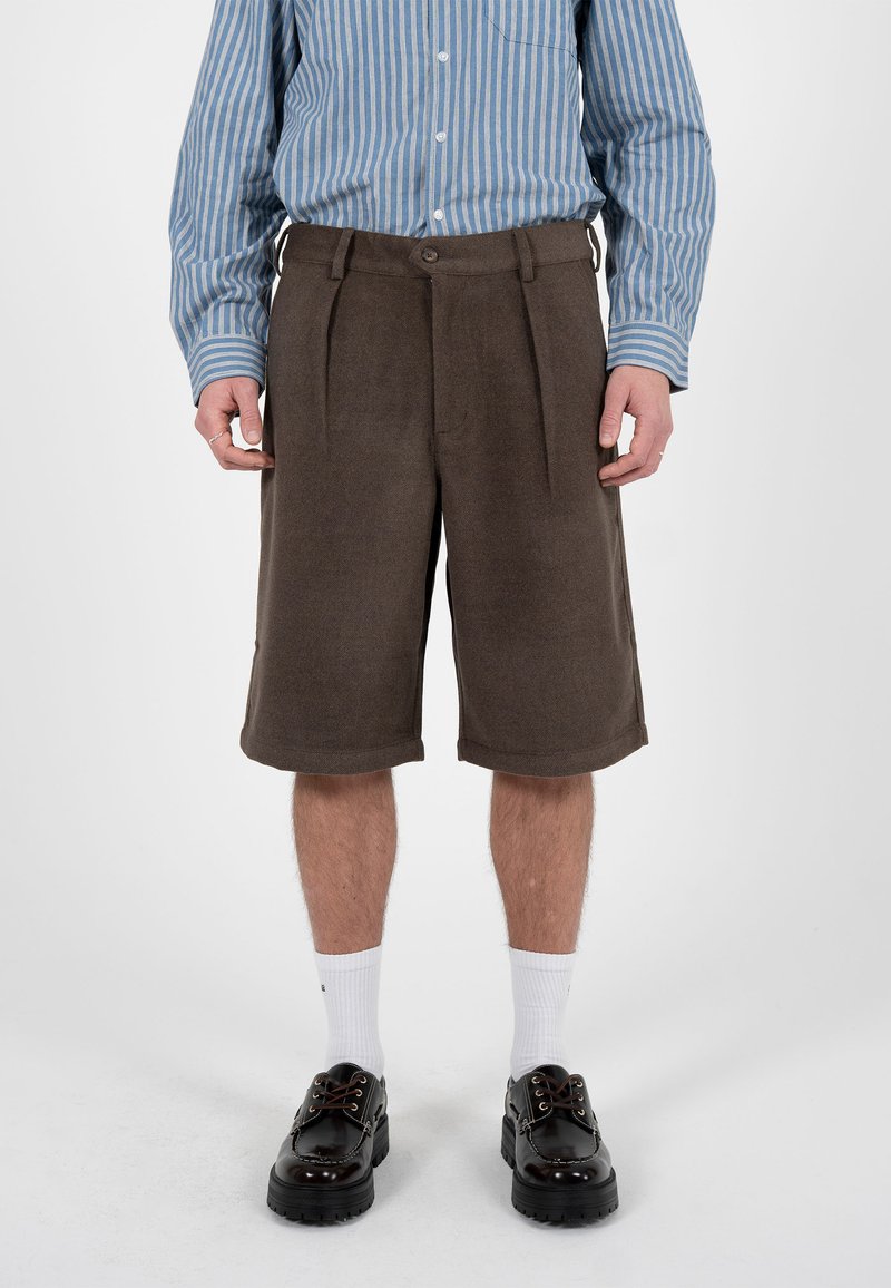 Homme portant un short marron au genou, une chemise à manches longues rayée bleu et blanc, des chaussettes blanches et des chaussures noires épaisses, debout sur un sol blanc.