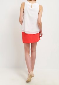 White sleeveless blouse with a keyhole back detail paired with a bright red skirt, finished with beige high heels. Smooth textures.