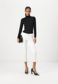 Maglione nero aderente a collo alto, jeans bianchi cropped con orlo ricamato e tacchi neri a punta. Borsa nera portata sul lato sinistro.