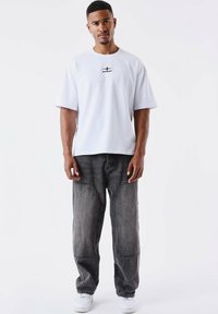T-shirt bianco in cotone con una piccola grafica nera al centro, abbinata a jeans denim grigi scuri a vestibilità rilassata e sneakers bianche.