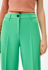 Kvinna som bär högmidjade ljusgröna byxor med en svart knapp, ihop med en kortärmad, ribbad orange topp.