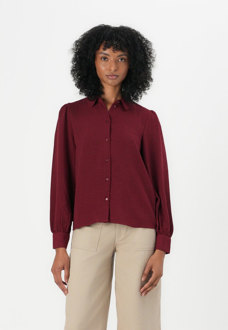 Cămașă burgundy cu butoni din material texturat, guler rotund, mâneci lungi bufante cu manșete ajustate și croială dreaptă. Asortată cu pantaloni bej.