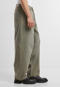 Baggy olive green pants made of cotton with side pockets and a label on the back. Worn with black footwear and a fitted white top.