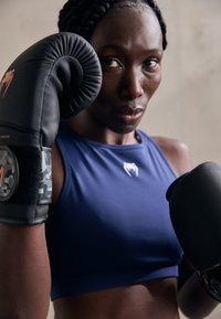Guantes de boxeo en negro con acentos rojos, usados por una persona con una camiseta sin mangas azul. Patrón de camuflaje notable en las correas de los guantes.