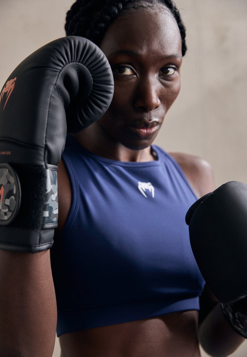 Guantes de boxeo en negro con acentos rojos, usados por una persona con una camiseta sin mangas azul. Patrón de camuflaje notable en las correas de los guantes.