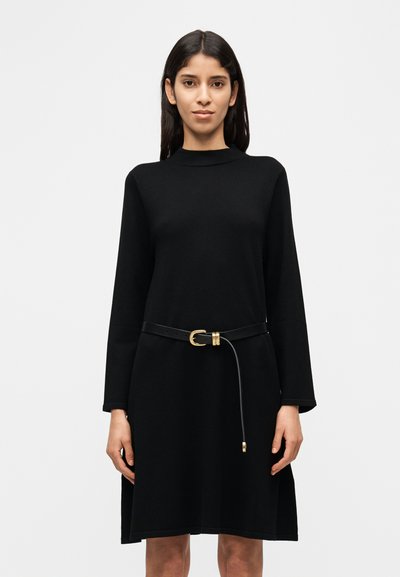 Robe noire à manches longues en tissu doux, avec un col montant et une ceinture noire fine à boucle dorée à la taille.