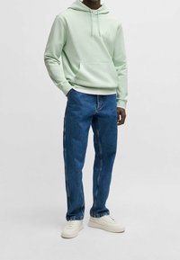 Mátově zelená mikina s kapucí na stažení a přední kapsou, v kombinaci s modrými džínami s postranními švy. Outfit doplňují bílé tenisky.