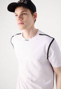 Fehér, rövid ujjú póló fekete részletekkel, kerek nyakkivágással és "BOSS" logóval a gallérnál. Sima pamutszövet.