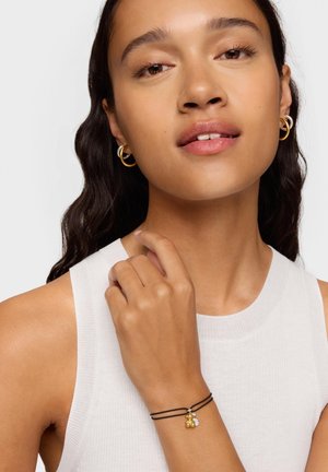 Pulsera de dijes de oro con dos dijes en cuerdas negras, llevada en una mano. La modelo tiene el cabello oscuro, lleva una blusa blanca sin mangas y pendientes de aro dorados.