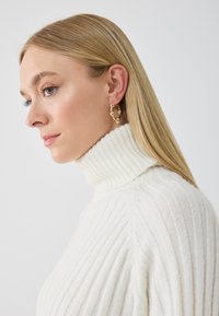 Suéter blanco de cuello alto con textura volumétrica. Pendientes de aro dorados con acentos y detalles circulares. Modelo mostrado de perfil.