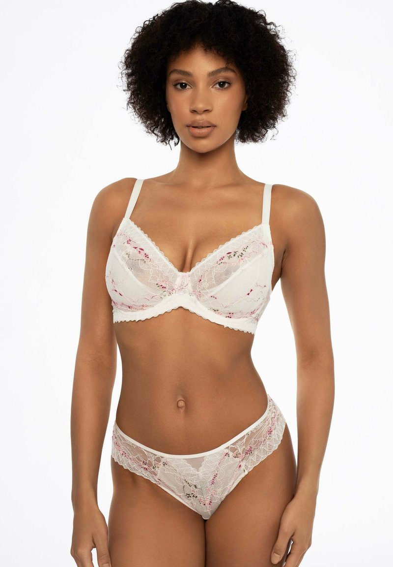 Koronkowy komplet bielizny w kolorze kości słoniowej, z różowymi kwiatowymi akcentami. Białe biustonosze z fiszbinami i falbaniastymi krawędziami; pasujące stringi dopełniają cały wygląd.