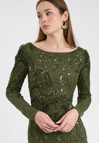 Abito a maniche lunghe di colore verde oliva con dettagli floreali in perline e paillettes. Presenta un colletto a barca e una silhouette aderente.