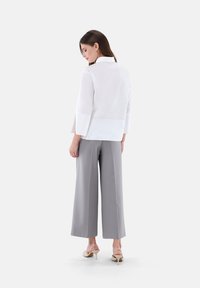 Weiße Bluse mit langen Ärmeln, hohem Kragen und seitlichen Schlitzen, kombiniert mit grauen weit geschnittenen Hosen und hellen Absatzsandalen.