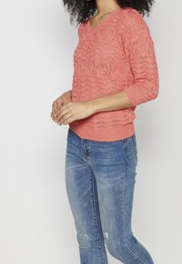 Pull en crochet corail avec un col en V, des manches ¾ et un motif texturé. Assorti à un jean denim bleu ajusté avec un effet usé.
