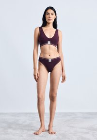 Burgundy urheilu bralette, jossa on V-päinen kaula-aukko ja elastinen vyötärö, yhdistettynä yhteensopiviin bikinin alaosiin, joissa on logotagi ja kontrastiväriset reunat.