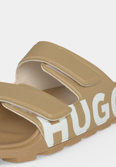 Sandale beige à enfiler avec deux brides réglables en velcro et nom partiel de marque blanc embossé sur le côté.