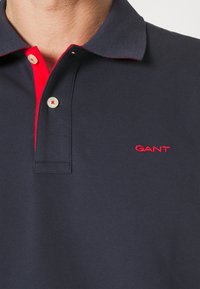 Tmavomodré polo s červeným golierom a légou so dvoma gombíkmi. Logo „GANT“ vyšitý červenou niťou na ľavej strane. Textúrovaná látka.