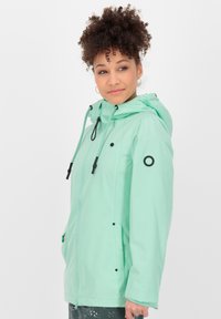 Veste vert menthe avec capuche, fermeture éclair, cordons de serrage et accents noirs à pression. Texture lisse, avec poches latérales et détail de logo.