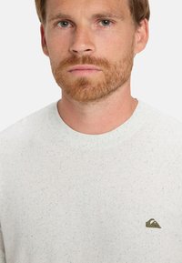 Lys grå bomulds t-shirt med rund halsudskæring, der har en spættet tekstur og et lille broderet logo i venstre brystområde.