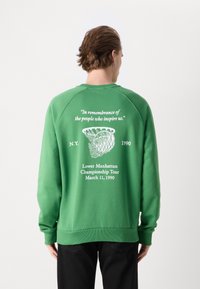 Grön sweatshirt med tryck på baksidan som har texten: "Till minne av de människor som inspirerar oss," och grafik, daterad 1990, rundtur i nedre Manhattan.