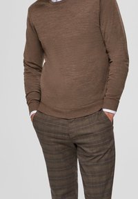 Brauner Pullover mit langen Ärmeln und ribbeltem Kragen, kombiniert mit strukturierten, karomusternden braunen Hosen. Beide Kleidungsstücke weisen ein lässiges Design auf.