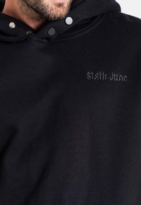 Felpa nera realizzata in tessuto misto cotone, con cappuccio regolabile tramite cordoncino, bottoni a pressione e ricamo frontale con la scritta "Sixth June" in filo scuro.
