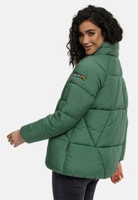 Groene pufferjack met een hoge kraag, geometrische stiksels en een kleurrijke patch op de mouw. Het materiaal lijkt glad en duurzaam.