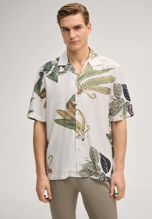 Junger Mann trägt beige Hosen und ein weißes kurzärmeliges Hemd mit grünen und braunen botanischen Blattmustern, steht vor einem schlichten Hintergrund.